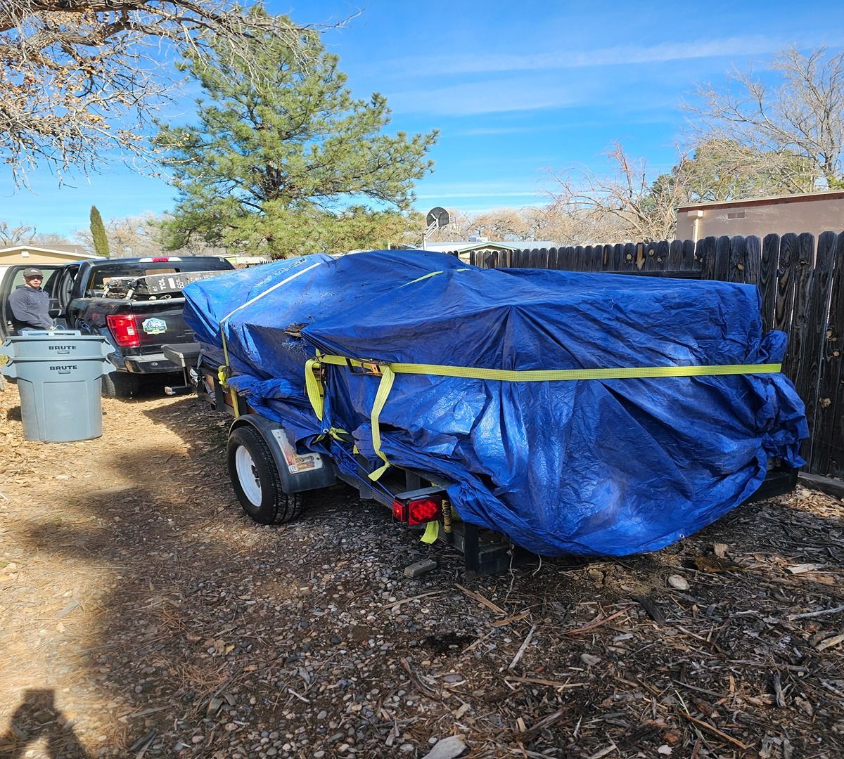 Blue tarp-covered trailer parked in a leaf-covered yard beside a black fence and trash bin.