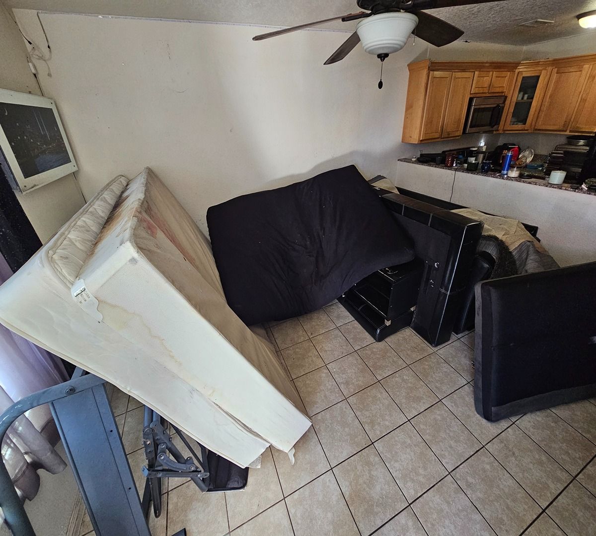 Cluttered living room with a tipped-over mattress, scattered furniture, tiled floor, and kitchen area in back