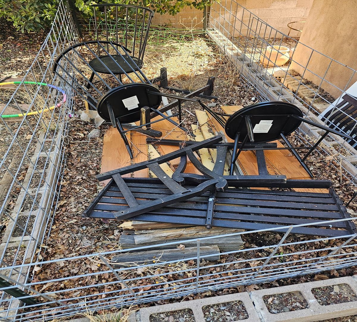 Two black outdoor chairs toppled on a metal patio bench in a debris-strewn fenced area