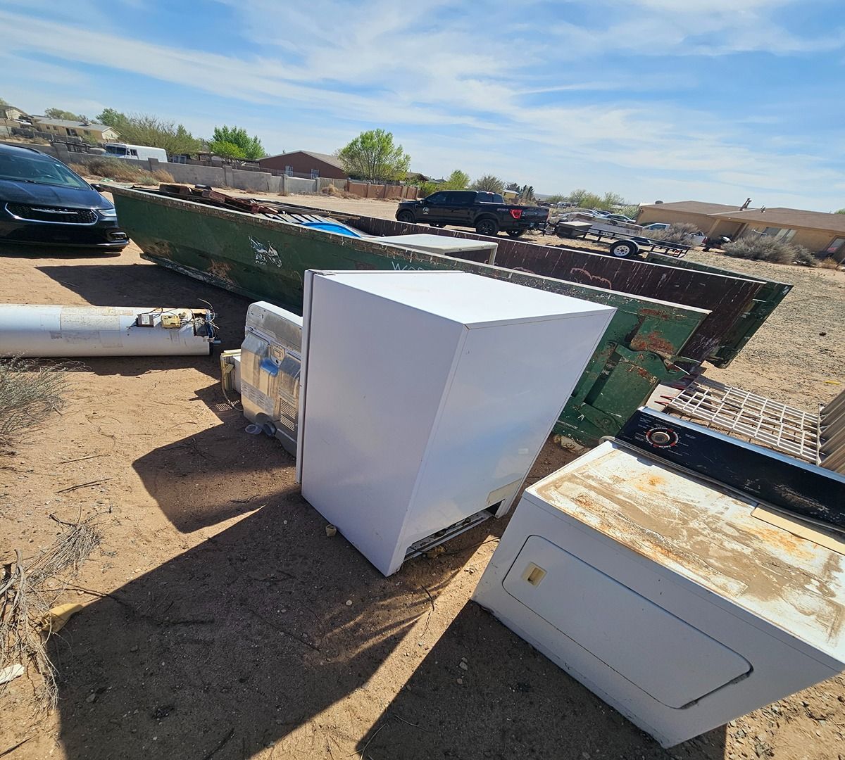 Discarded appliances in a dusty lot, including a white refrigerator and freezer near a dumpster.