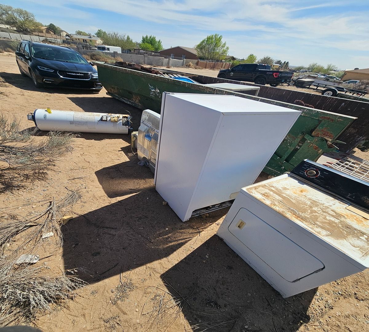 Discarded appliances and a car in a dusty yard under bright sun