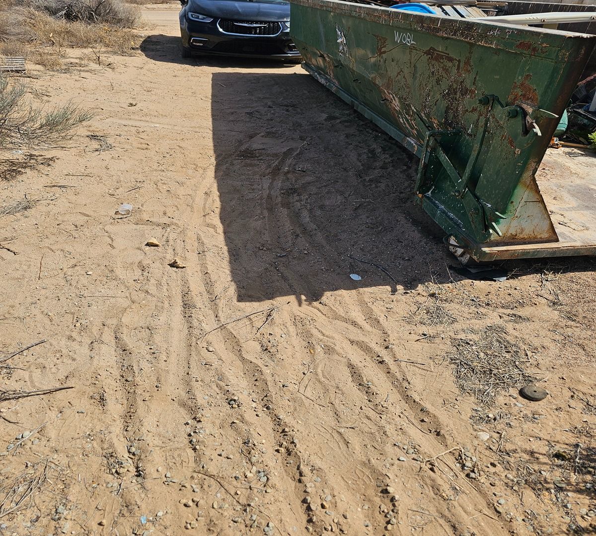 A dusty path with a black car and a large green metal dumpster blocking part of the road.