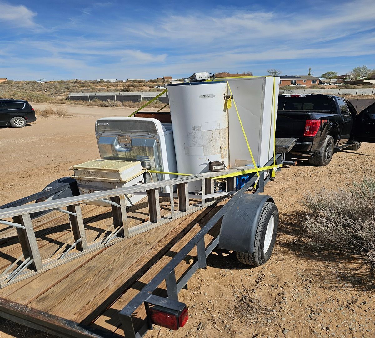Utility trailer loaded with white tanks and lumber on a dirt lot, with a black pickup nearby