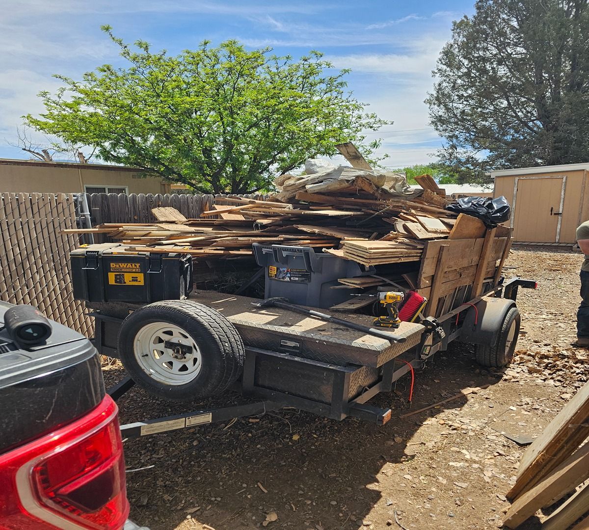 Utility trailer piled with lumber in a sunny dirt lot, with a red vehicle edge in the foreground