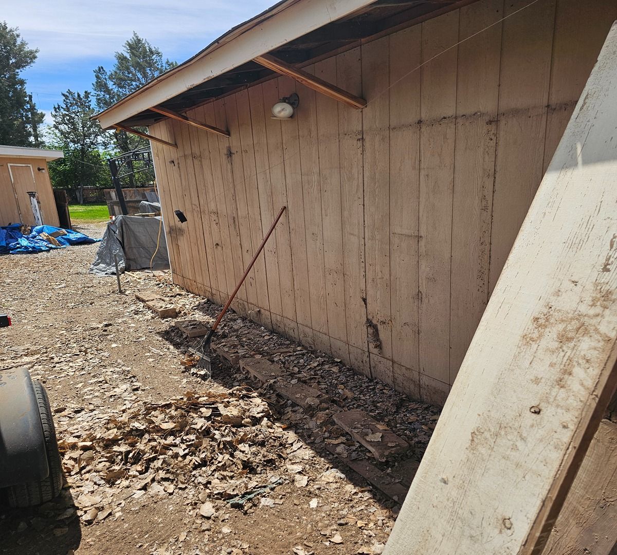 Side of a tan building with debris-strewn dirt ground and a large board leaning against the wall