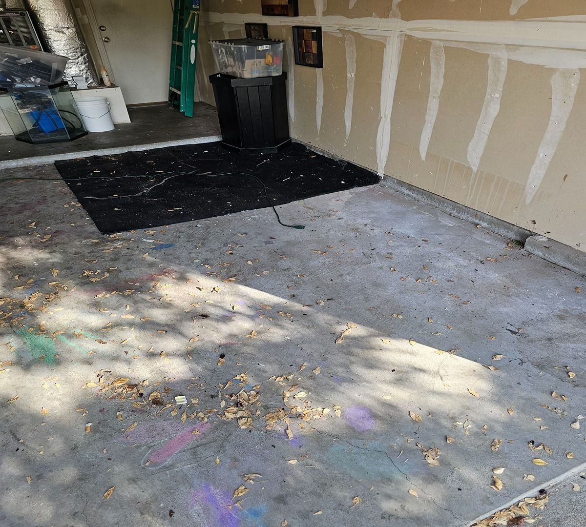 Garage floor with black mats, drywall patches, and scattered leaves on a concrete surface