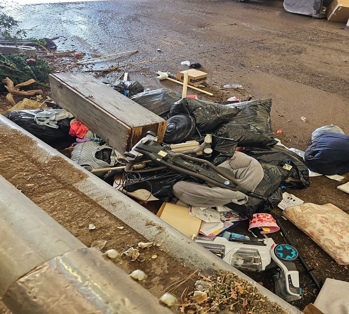 Pile of trash and debris beside a wet roadside curb, including broken items and cardboard.