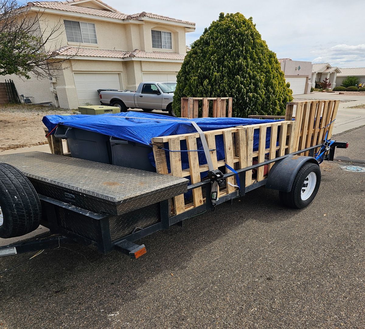 Flatbed trailer loaded with lumber, a blue tarp, and a large green shrub in a suburban driveway.