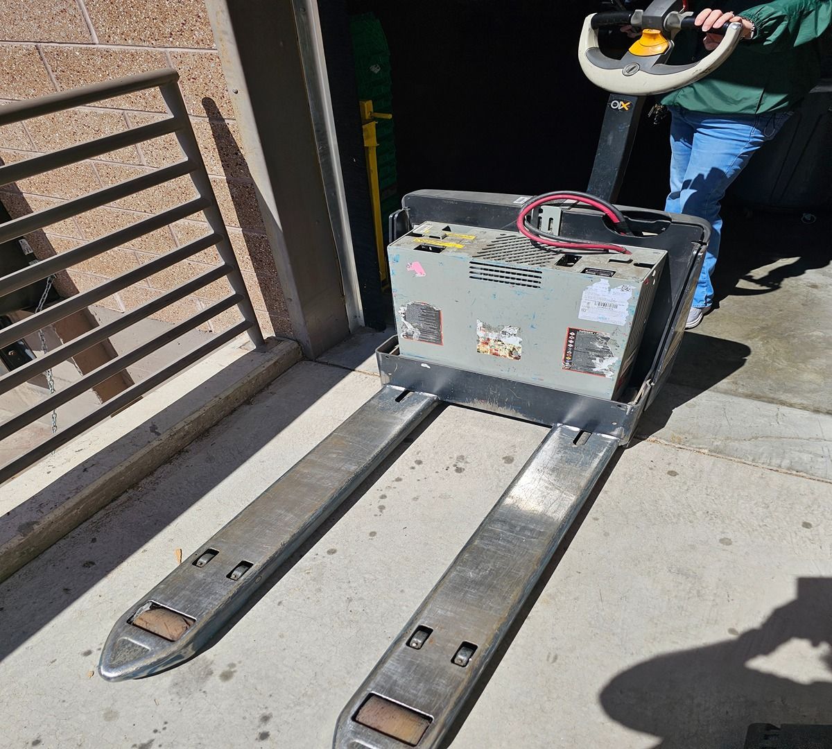 Worker operates a forklift with long forks beside a loading dock and metal gate.