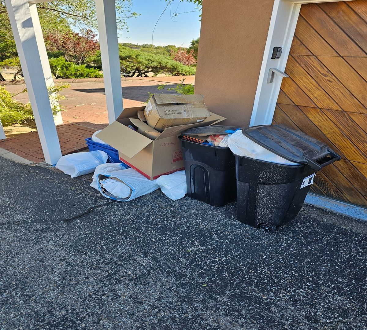 Trash bags and cardboard boxes piled by a porch beside a house entrance.