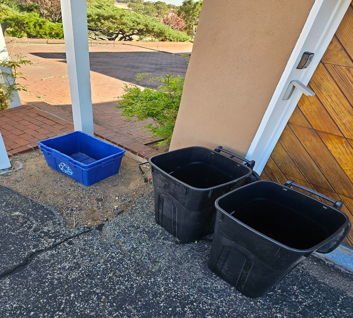 Three empty bins, two black and one blue, on a porch beside a post and garage door.