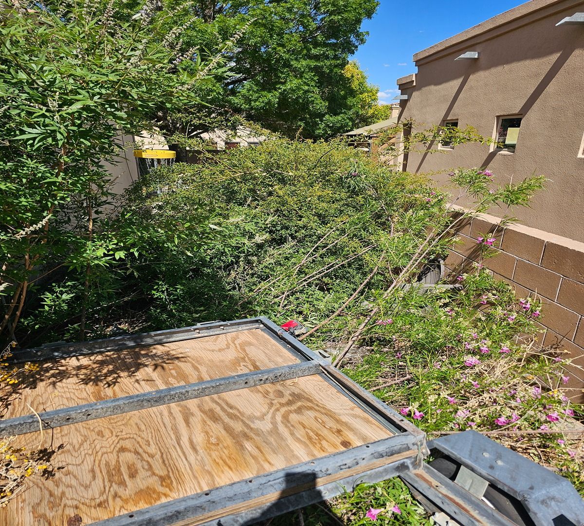 Wooden trailer or truck bed beside a house, surrounded by dense green bushes and flowers.