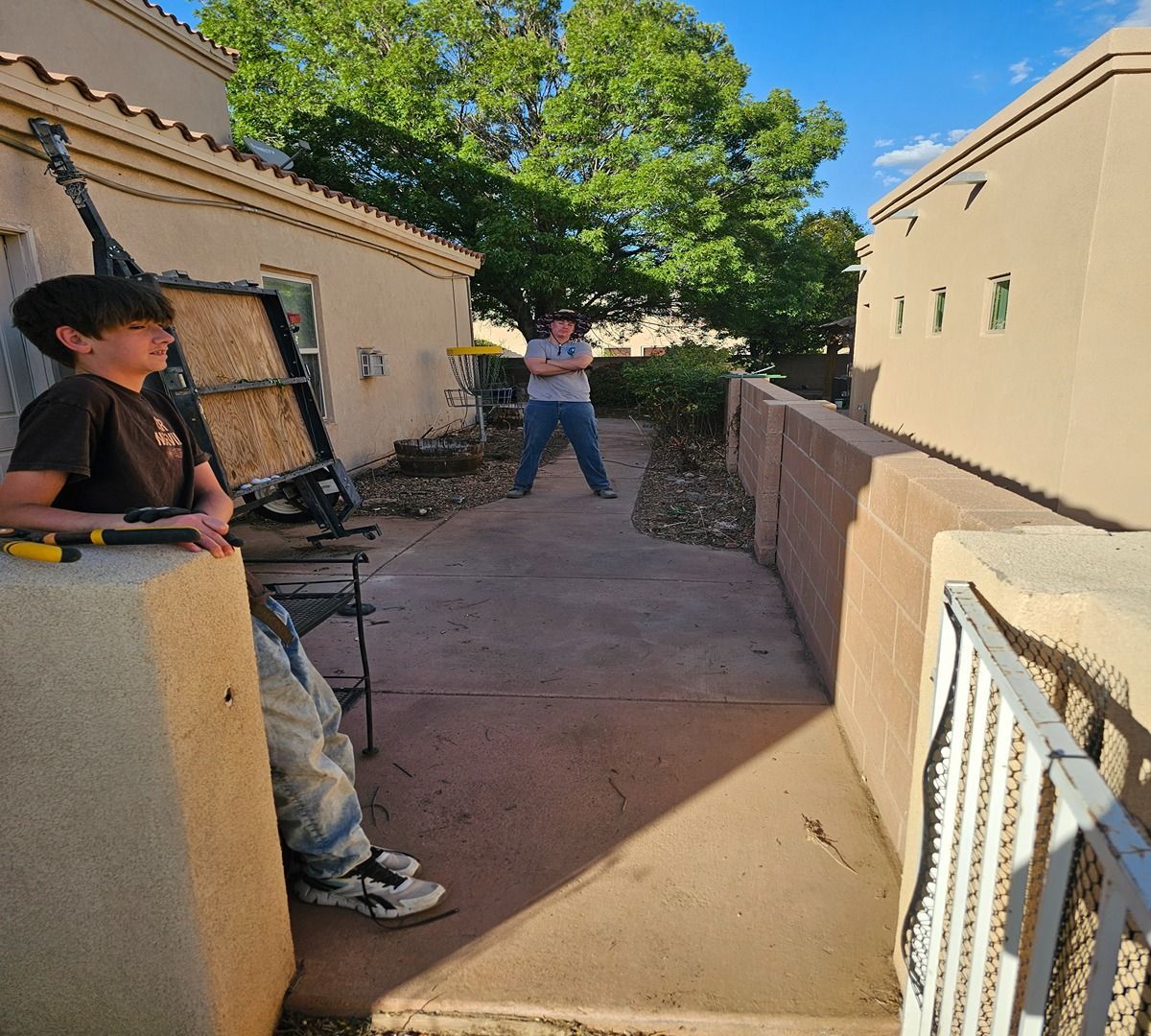 Child on the left and person in the distance in a sunlit outdoor courtyard with tan walls and trees.