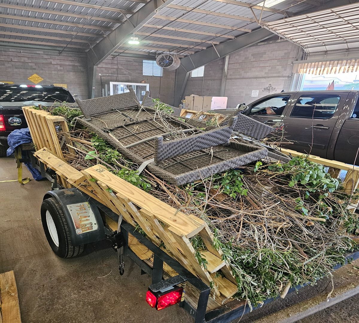 Trailer loaded with stacked wooden pallets and debris inside a barn-like garage, with a car nearby