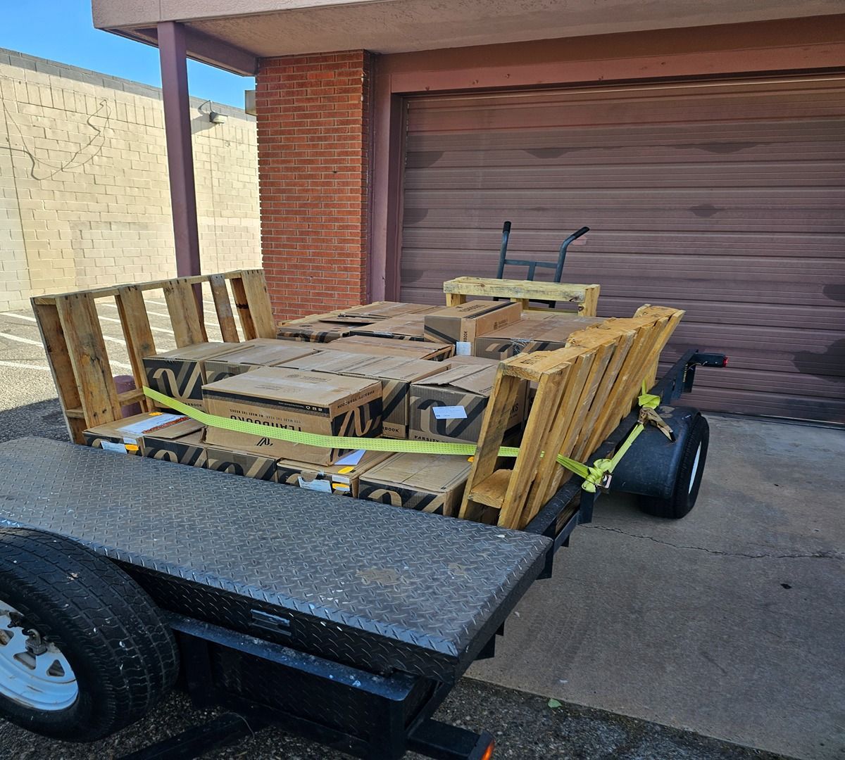 Flatbed trailer loaded with wooden furniture parts and straps, parked in front of a garage door.