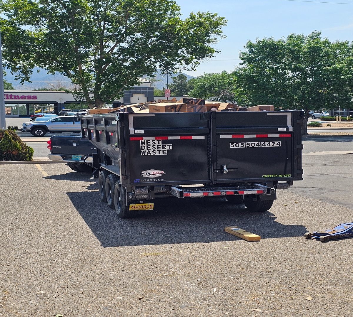 Black utility trailer loaded with wood, parked on gravel lot near trees and storefronts