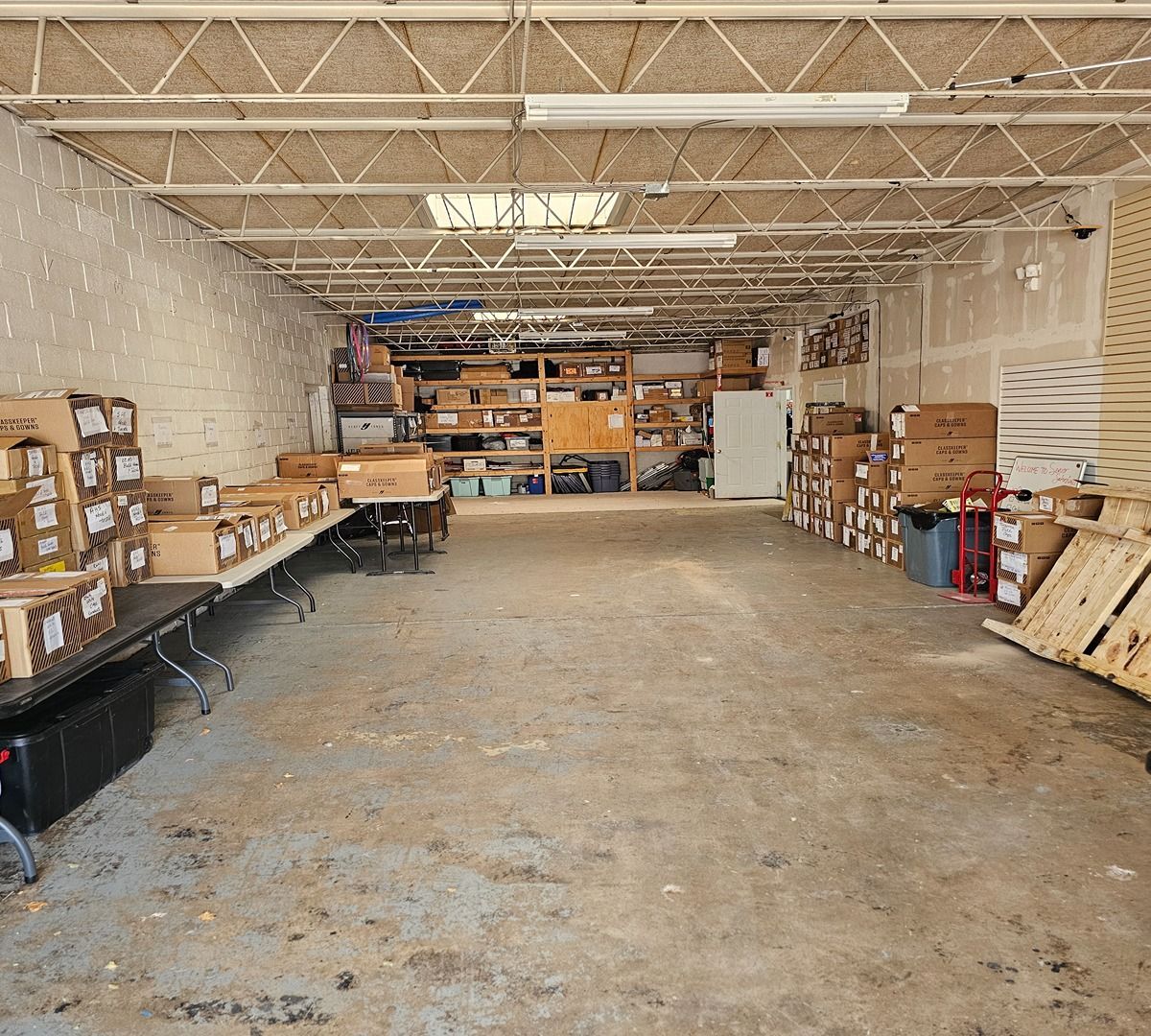 Warehouse interior with shelves, stacked boxes, and folding tables along a large open concrete floor.