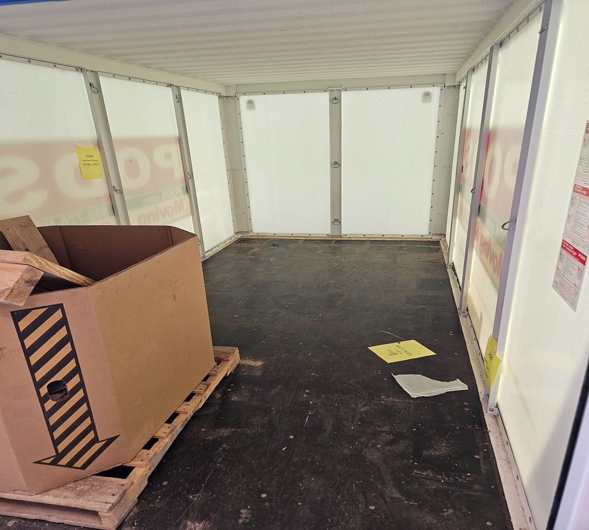 Empty shipping container interior with a palletized cardboard box on the left and papers on the floor.