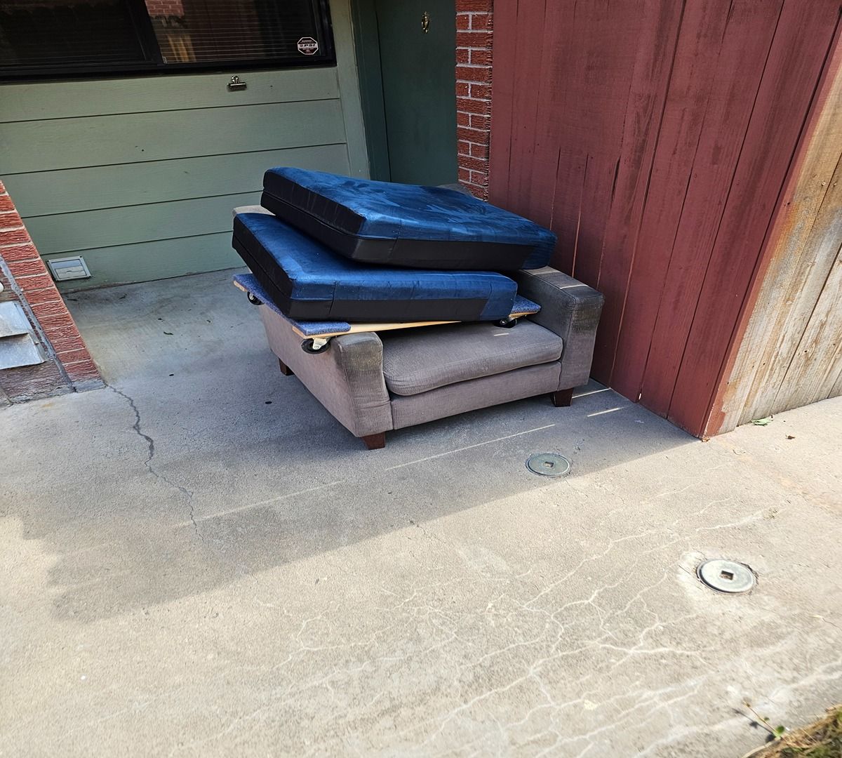 Stacked mattresses and a couch on a concrete walkway beside a house and red fence