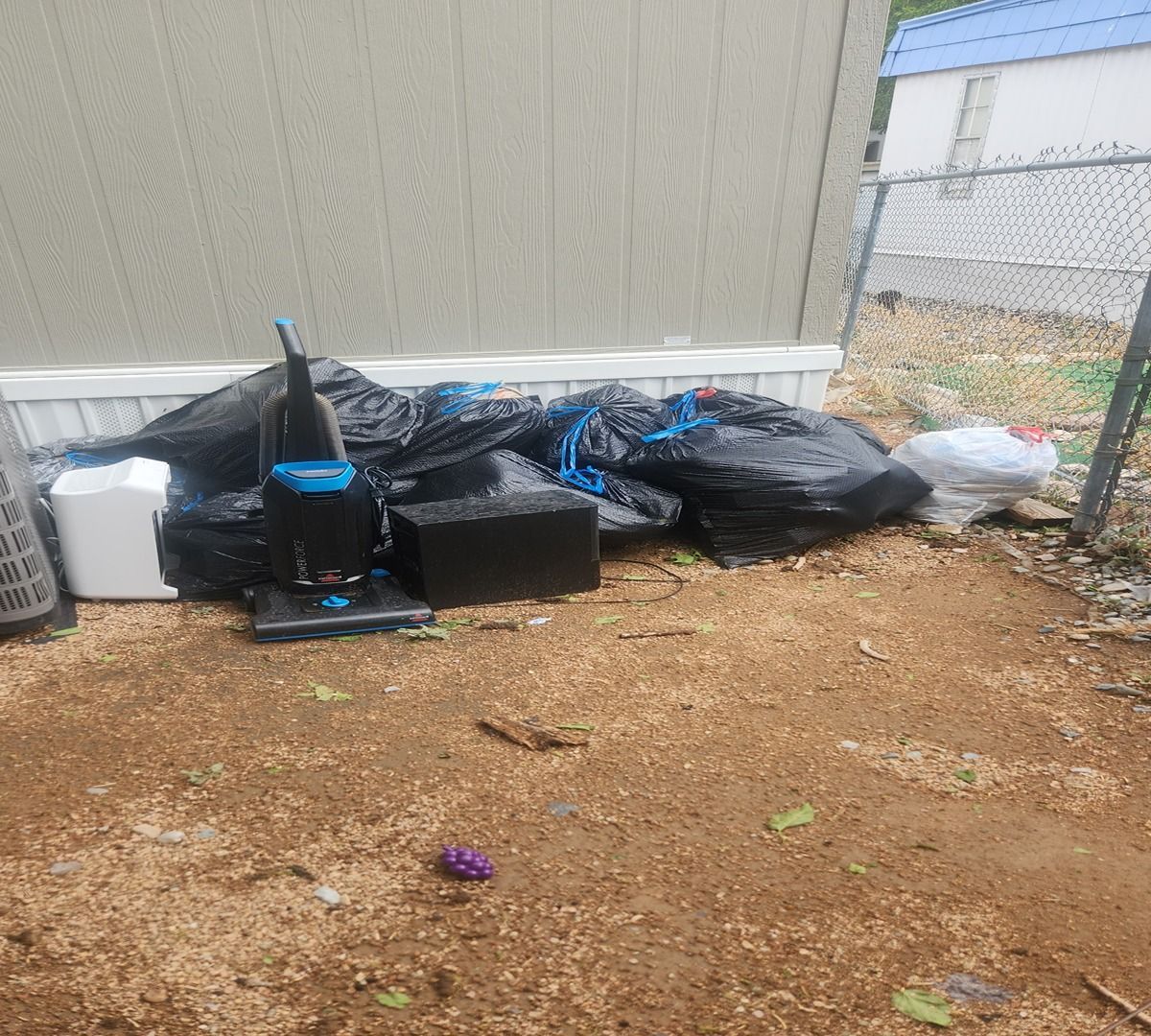 Trash bags and a cooler piled against a beige wall on a dirt patch beside a chain-link fence.