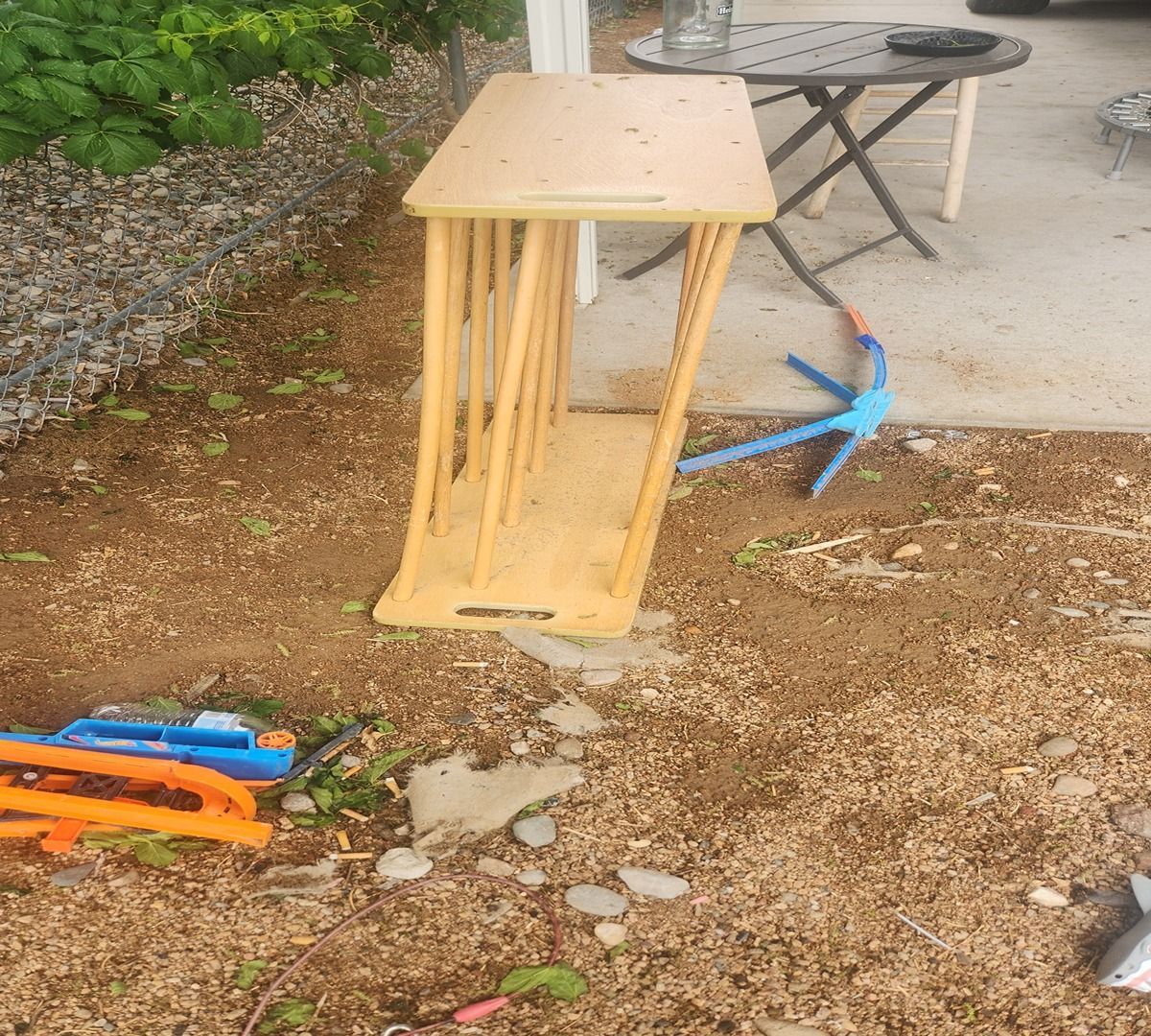 Overturned wooden side table on dirt beside a concrete patio, with a blue stand and colorful plastic pieces nearby