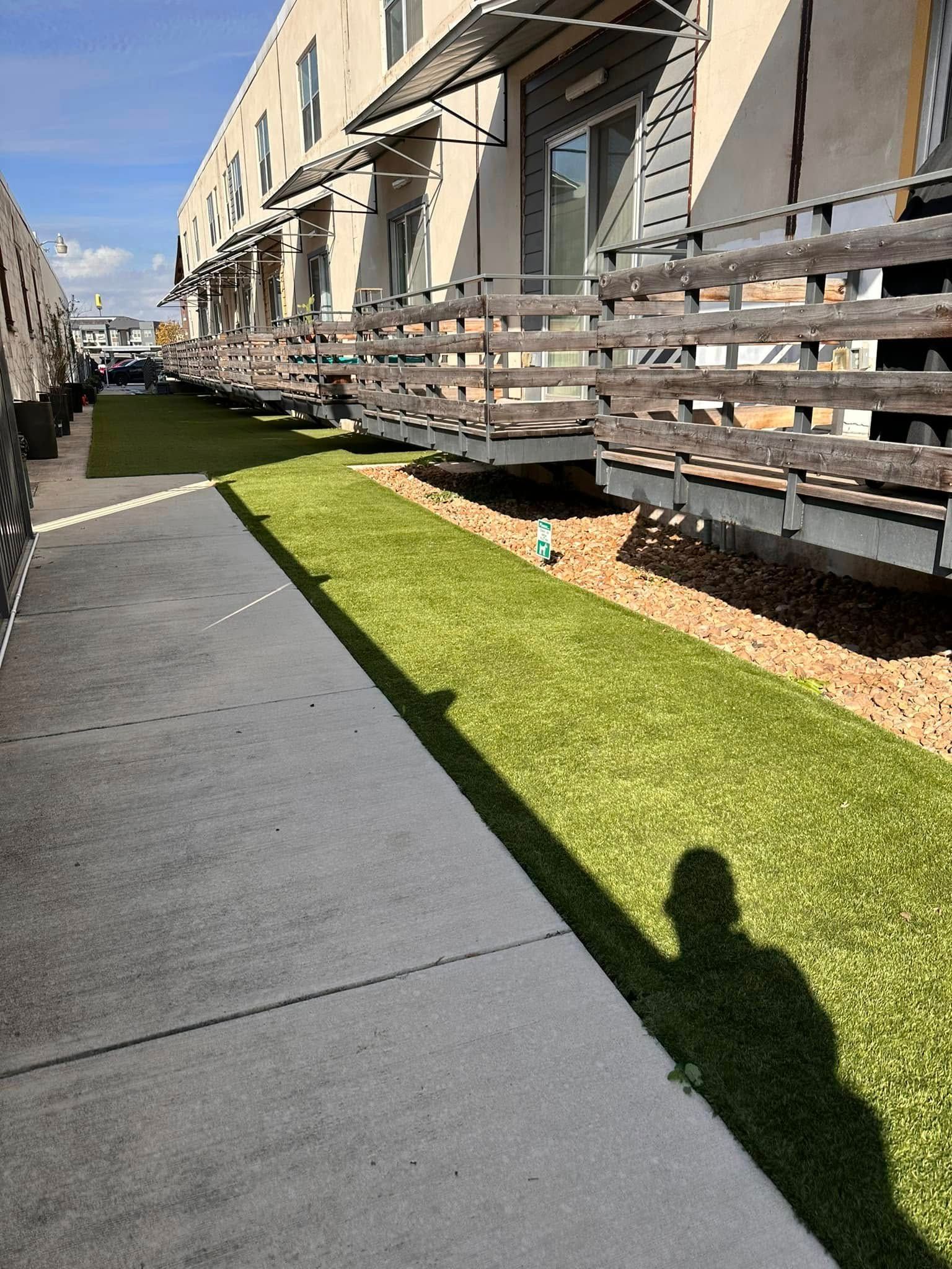 A person's shadow on a cement walkway beside artificial turf and a building with balconies.