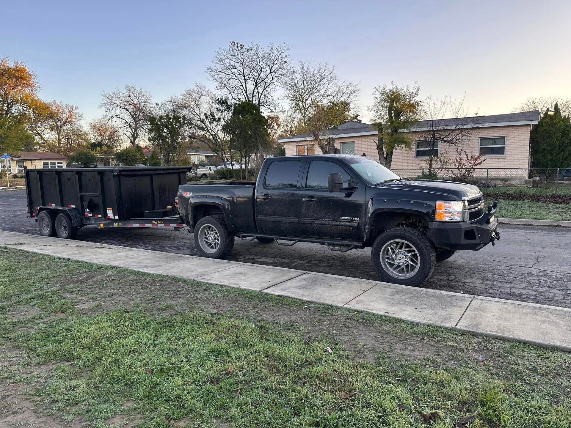 Black pickup truck towing a black dumpster trailer parked on a sidewalk next to a grassy area.