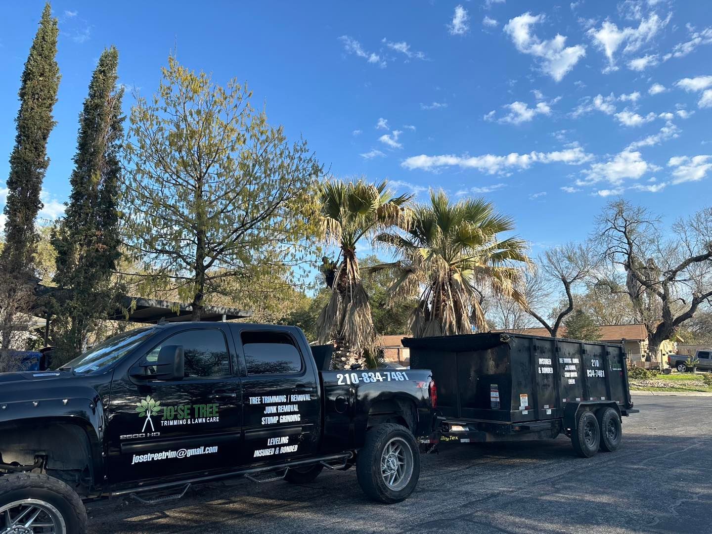Black truck towing a trailer with a dumpster, parked outdoors under a blue sky with trees.