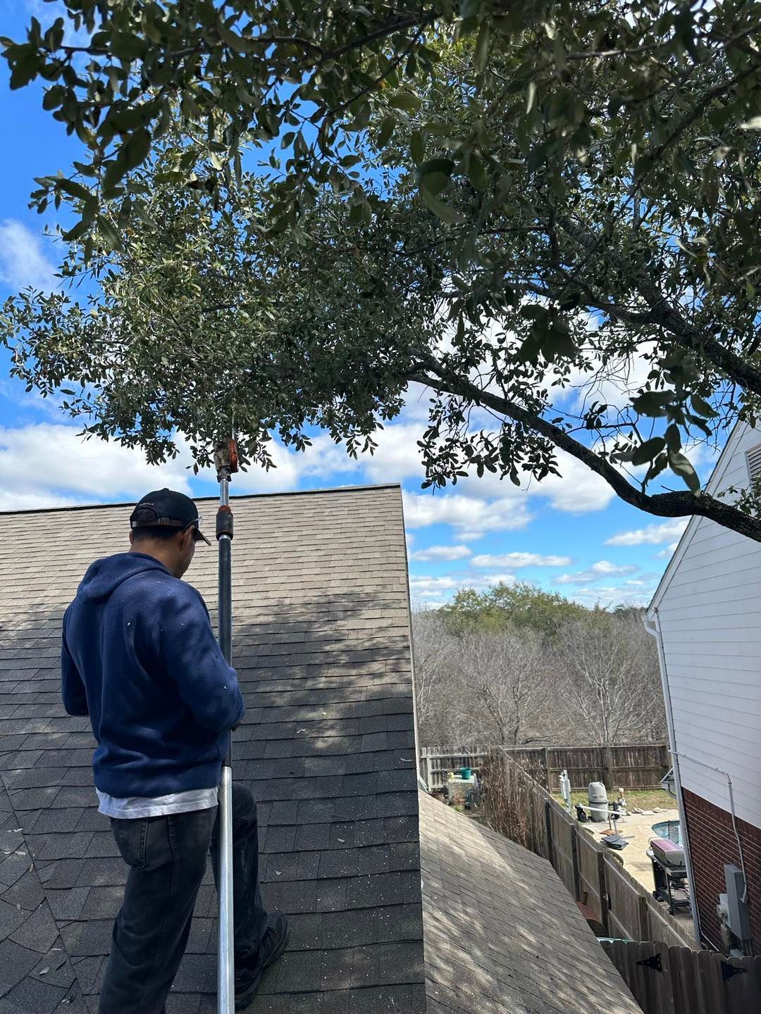 Person on a roof using a tool to trim tree branches. Blue sky with clouds and neighboring house.