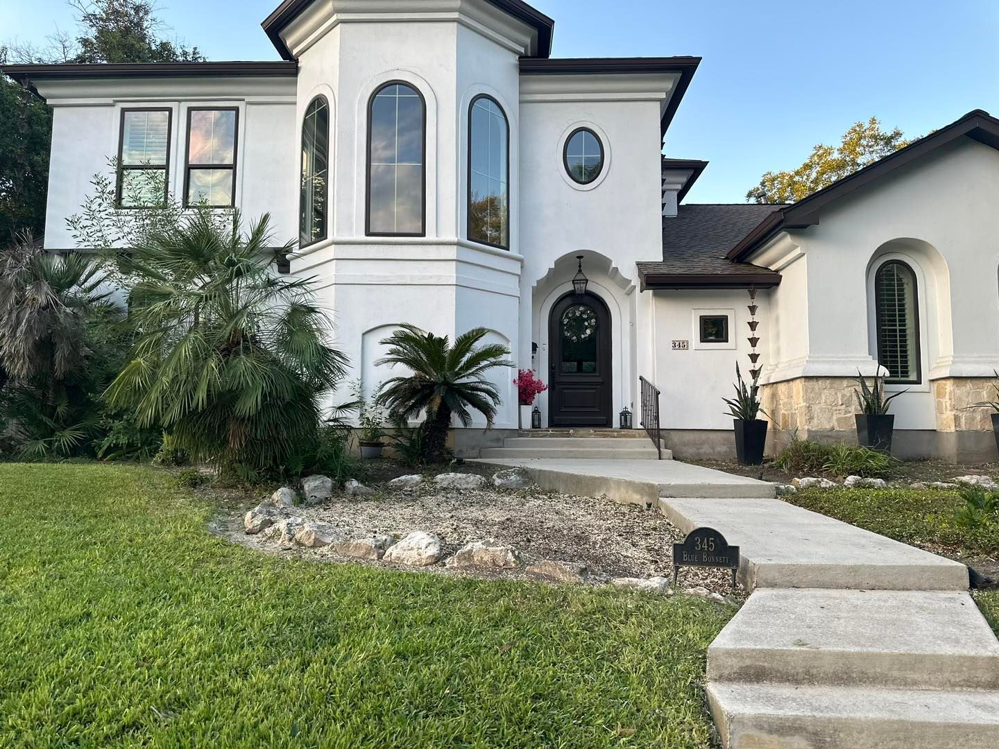 White two-story house with black trim, curved windows, a turret, and a stone-covered base.  A concrete pathway leads to the front door.
