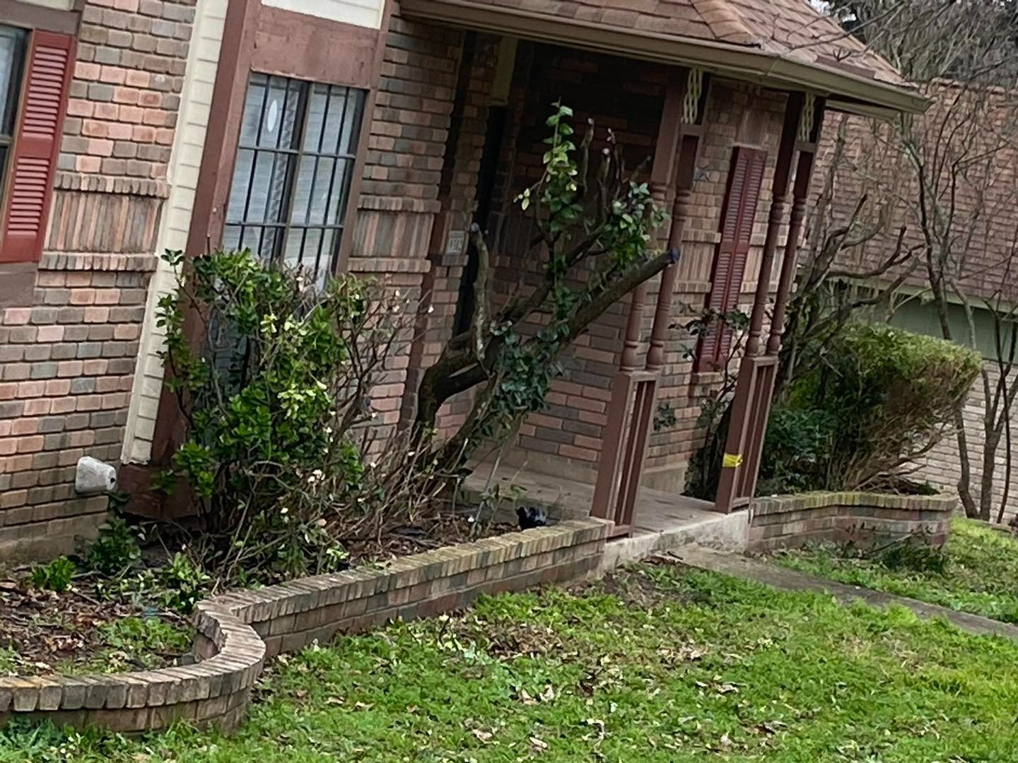 Brick house with porch, slanting slightly. Bushes and small trees surround the entrance.