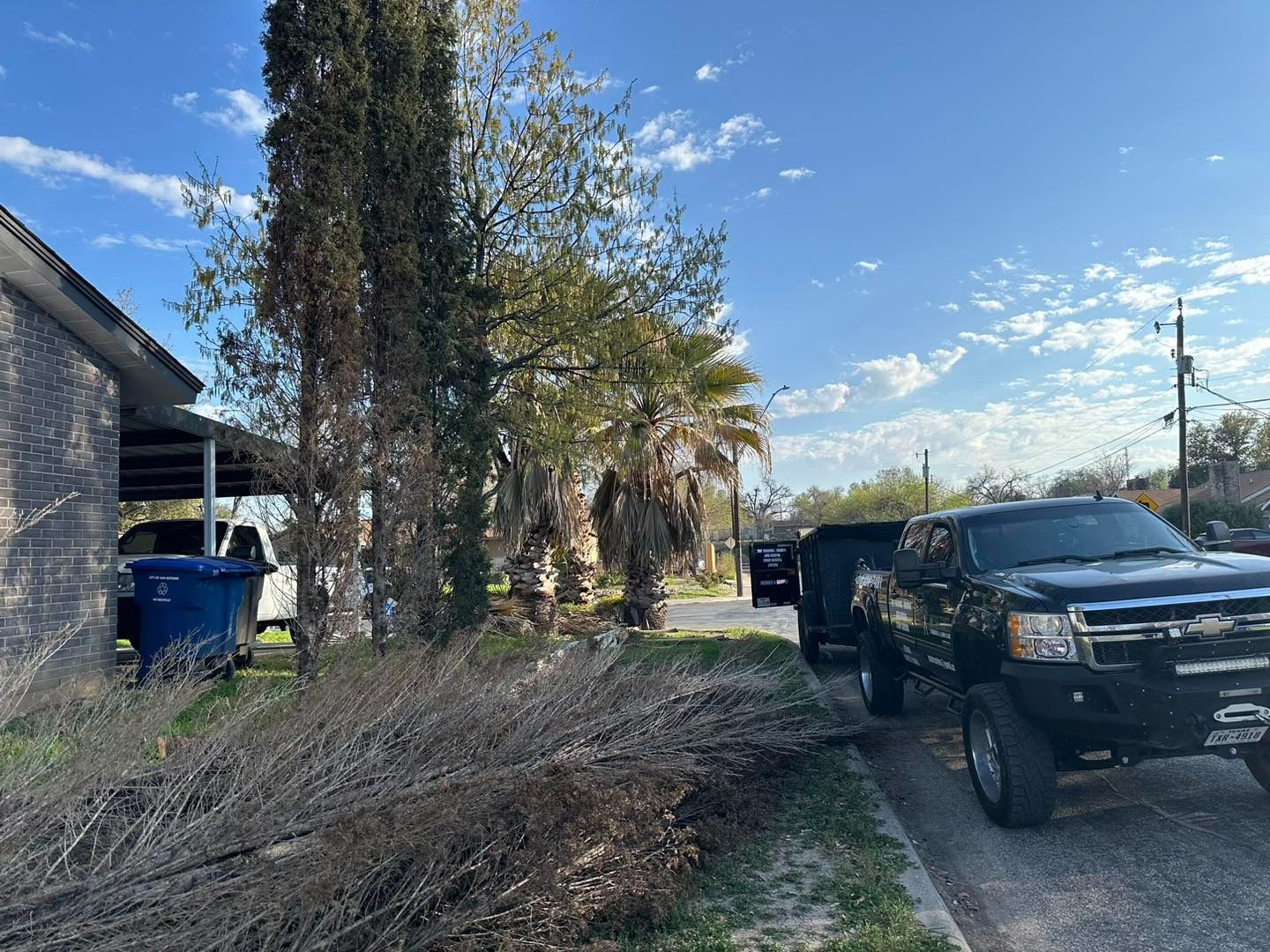 Black truck parked next to a trailer on a driveway. House and trees are in the background. Blue sky.