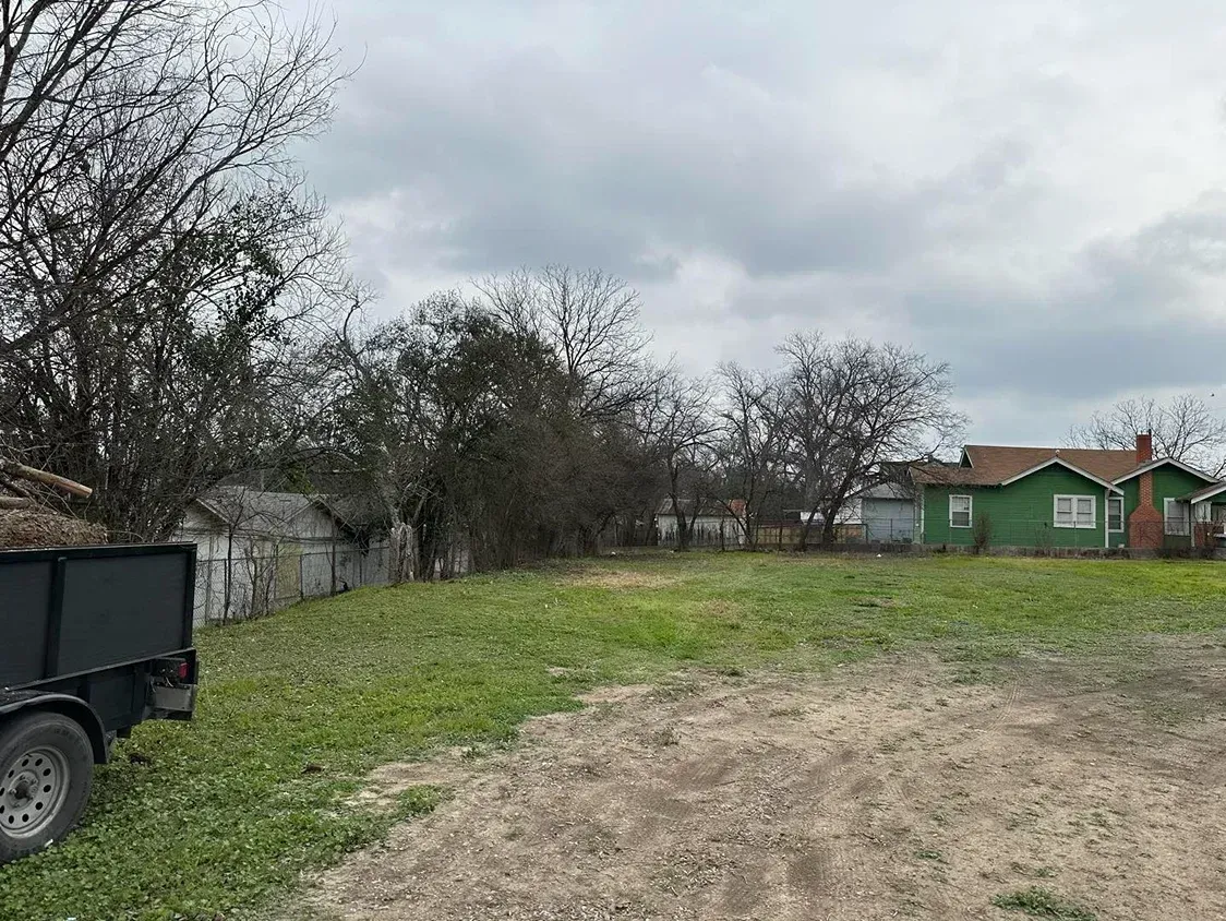 A vacant lot with a trailer, trees, and a green house under an overcast sky.