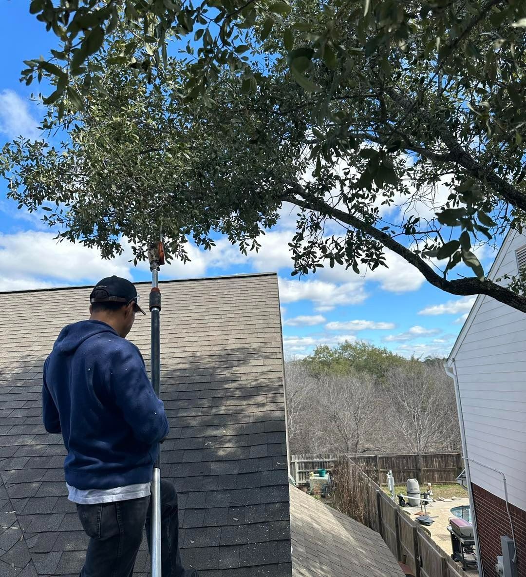 Person on a roof trims tree branches with a pole saw. Blue sky, white house visible.