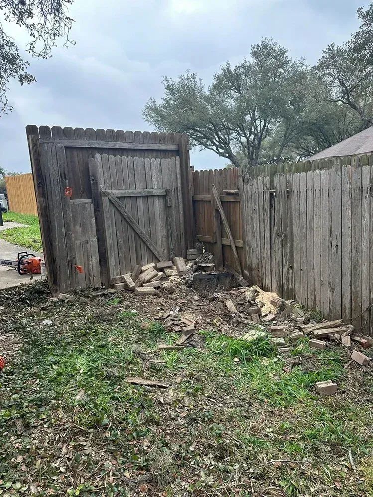 Damaged wooden fence with debris, small gate, and overgrown grass in a yard.