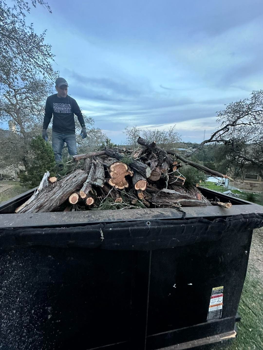 Man standing near a full dumpster of wood. Cloudy sky, outdoor setting.