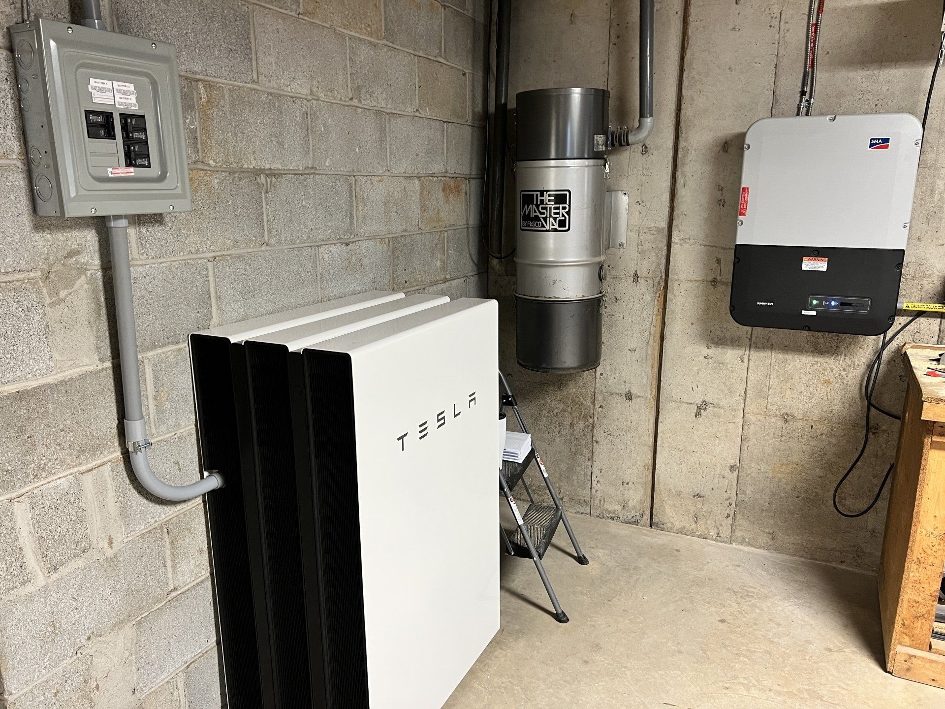 A tesla battery is sitting in a basement next to a vacuum cleaner.