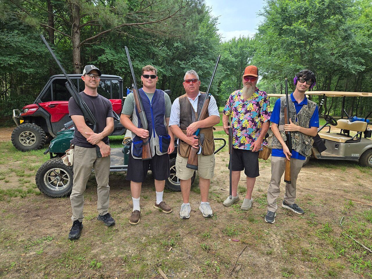 Five people pose with shotguns, golf cart, and ATV in a wooded area. They wear vests and look at the camera.