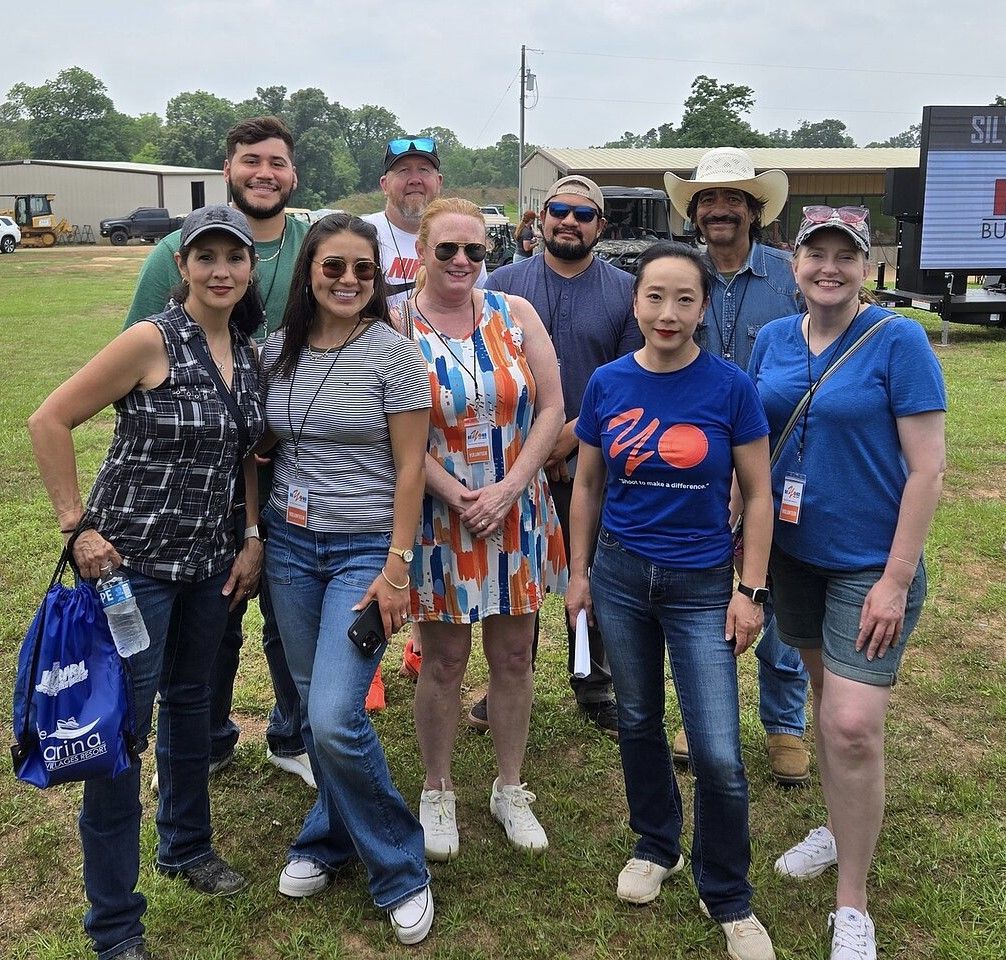Group of nine people smiling in an outdoor field. Some wearing lanyards, some in casual attire.