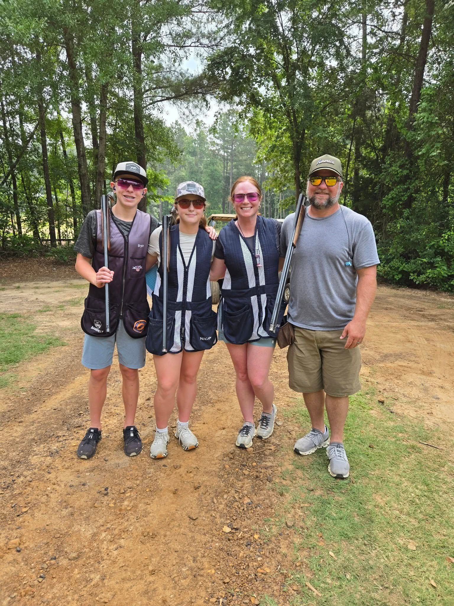 Four people with shotguns stand outside in front of trees. They wear vests, sunglasses, and shorts.