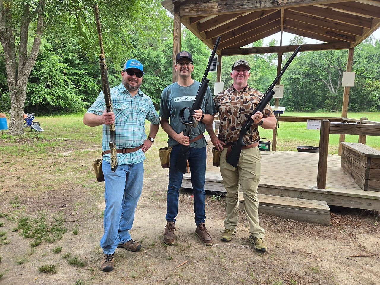 Three people at a shooting range, holding rifles. They pose smiling near a wooden structure.