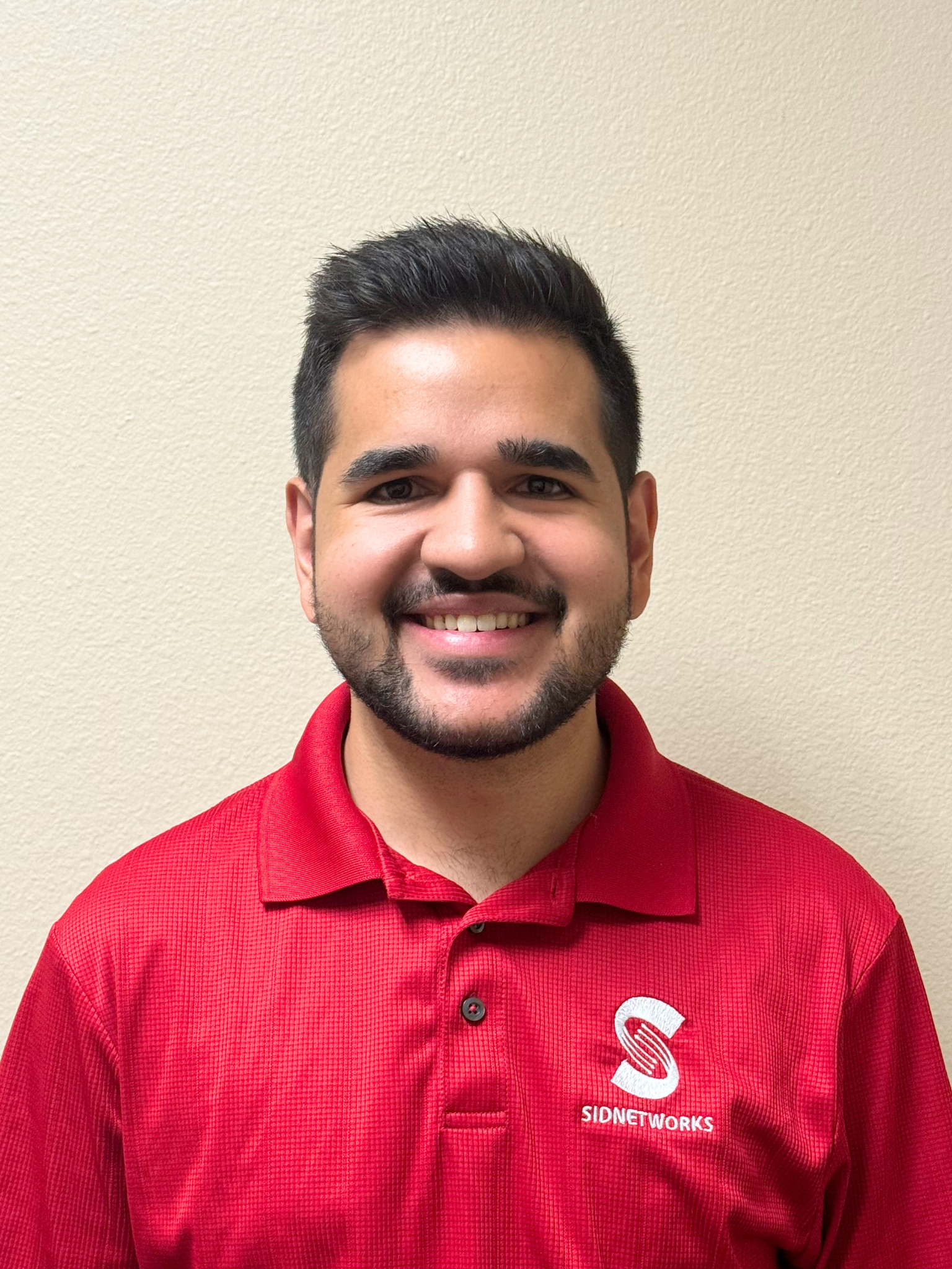 Man wearing a red polo shirt, smiling, with a logo on the left chest, in front of a plain wall.