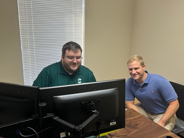 Two men smiling at a computer screen in an office. One in a green shirt, one in a blue polo.