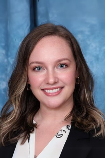 Woman smiling, wearing a black blazer and patterned blouse, in front of a blue background.