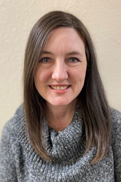 Woman with shoulder-length brown hair, smiling, wearing a gray sweater, standing in front of a neutral-colored wall.