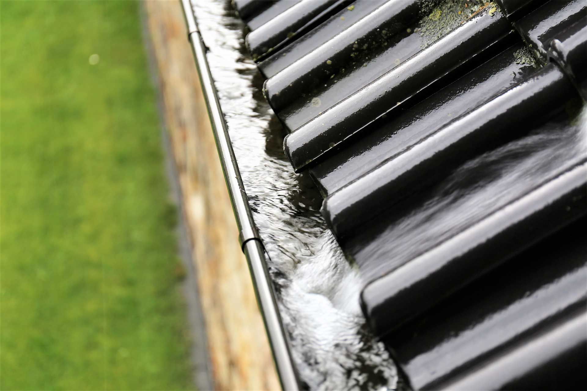 Rainwater flowing rapidly through a dark gutter on a roof, next to a green lawn.