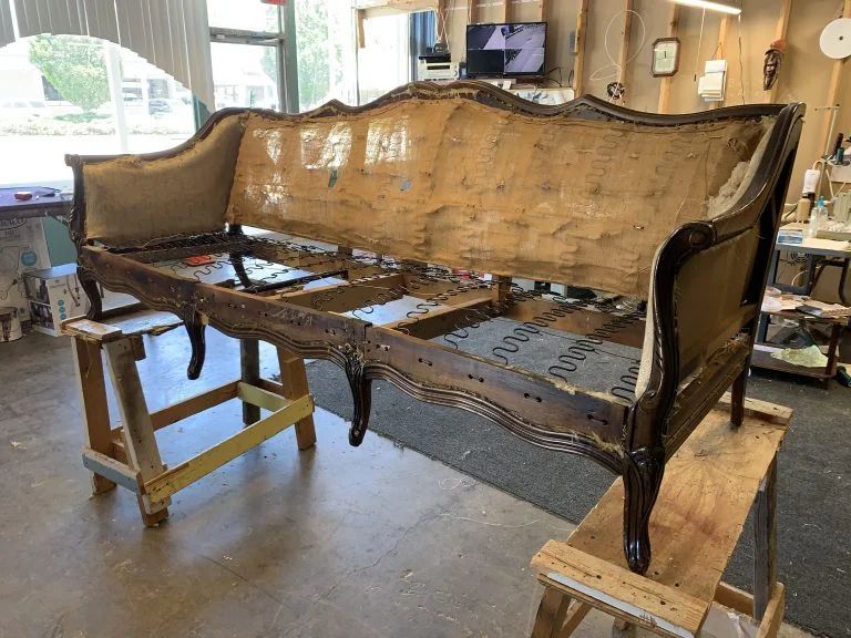 Damaged antique sofa frame in a workshop, with exposed wood and missing upholstery.