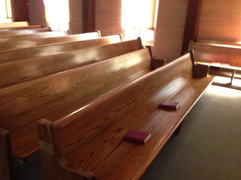Wooden church pews in a sunlit sanctuary, with hymnals on the seats.