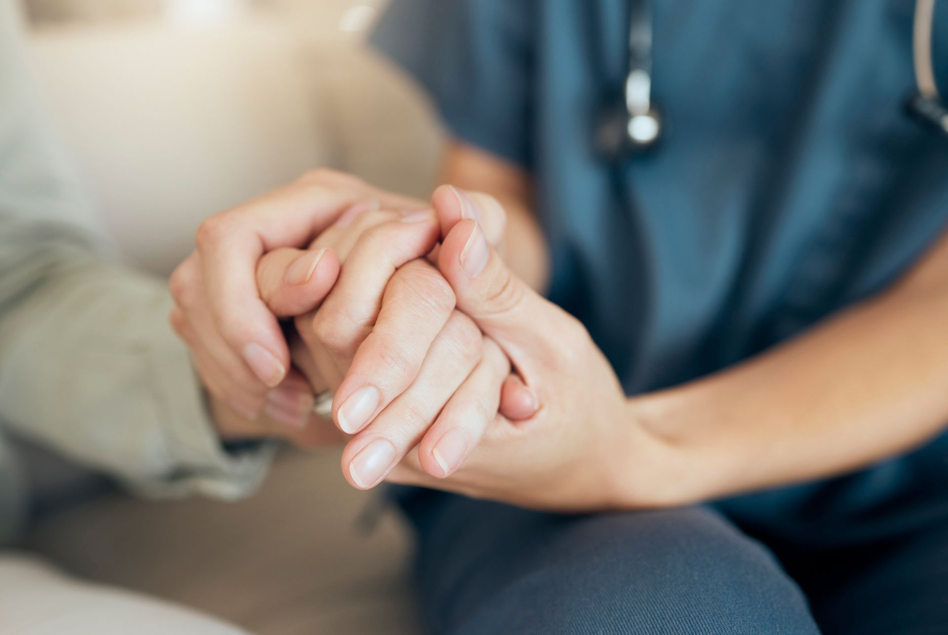 A medical professional in blue scrubs holding a patient’s hands in a gesture of comfort and support.
