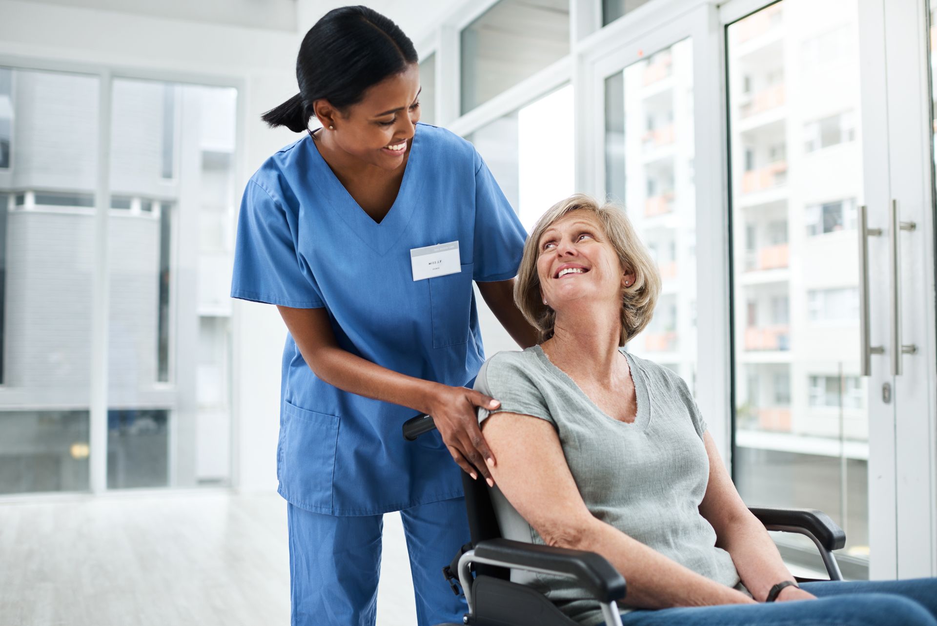 A healthcare professional in blue scrubs smiling as they assist a person in a wheelchair near a large window.